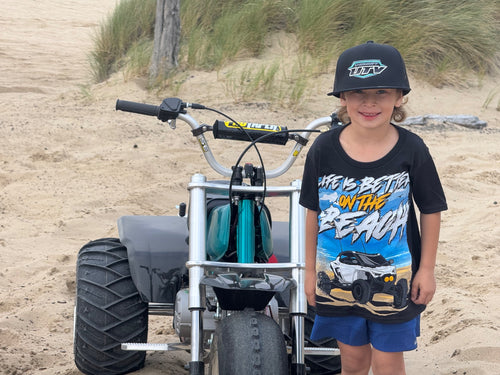 Child wearing black Showoff UTV Life Is Better On The Beach kids tee beside a mini bike in the dunes